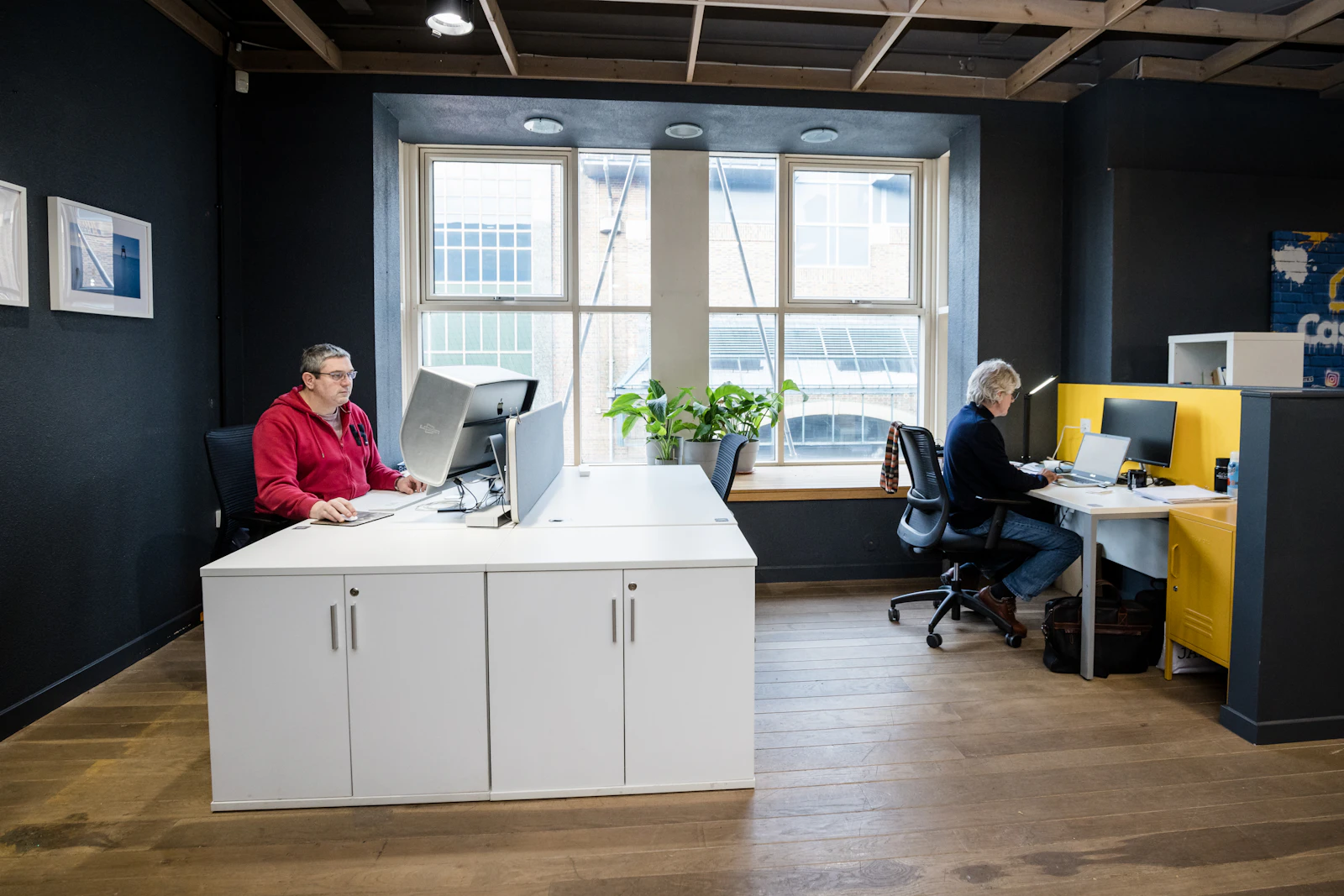 Two people working at desks in a bright shared office with large windows